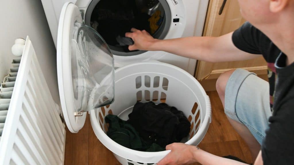 A man is kneeling while loading dark laundry from a white basket into a front-loading washing machine.