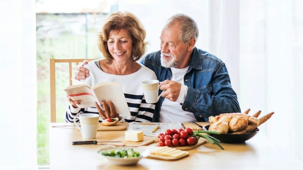 An older couple smiles at a table, with the woman reading a book and the man holding a mug.