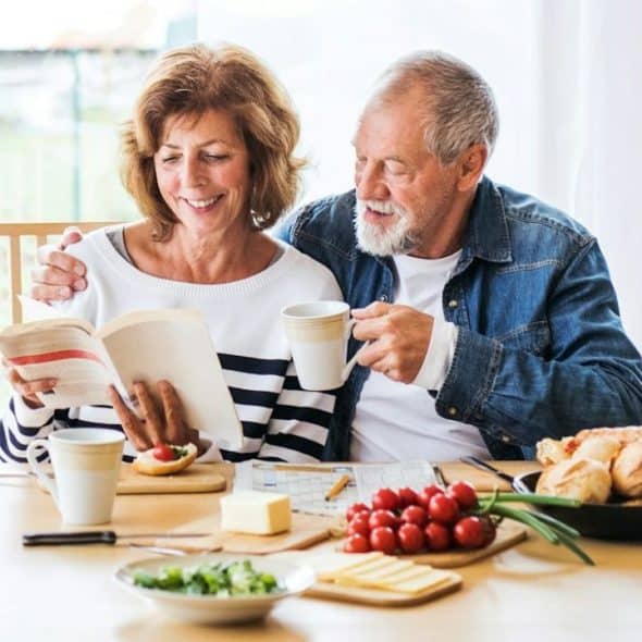 An older couple smiles at a table, with the woman reading a book and the man holding a mug.
