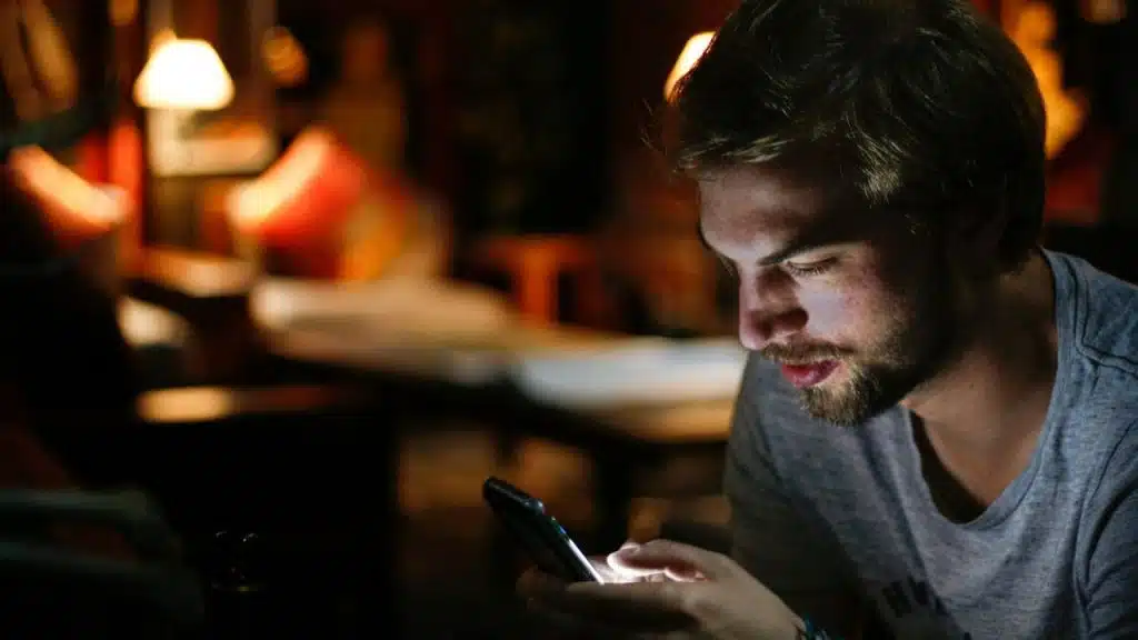 A young man with a beard sits in a dimly lit setting, looking down at his illuminated phone.