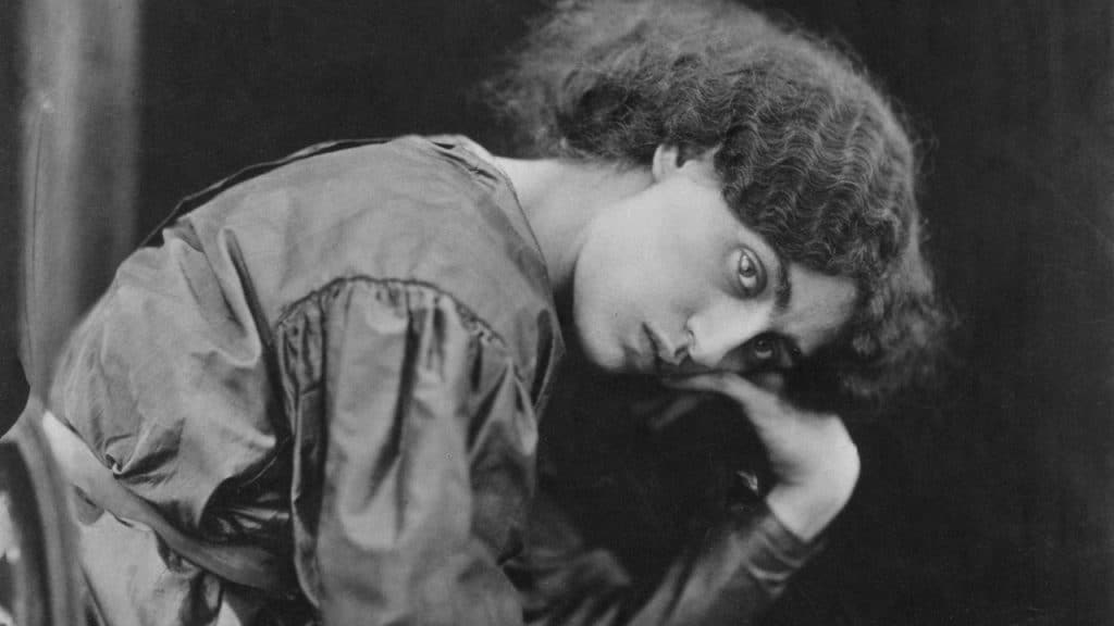 A black and white close-up photo of a woman with curly hair resting her chin on her hand and looking directly at the camera.
