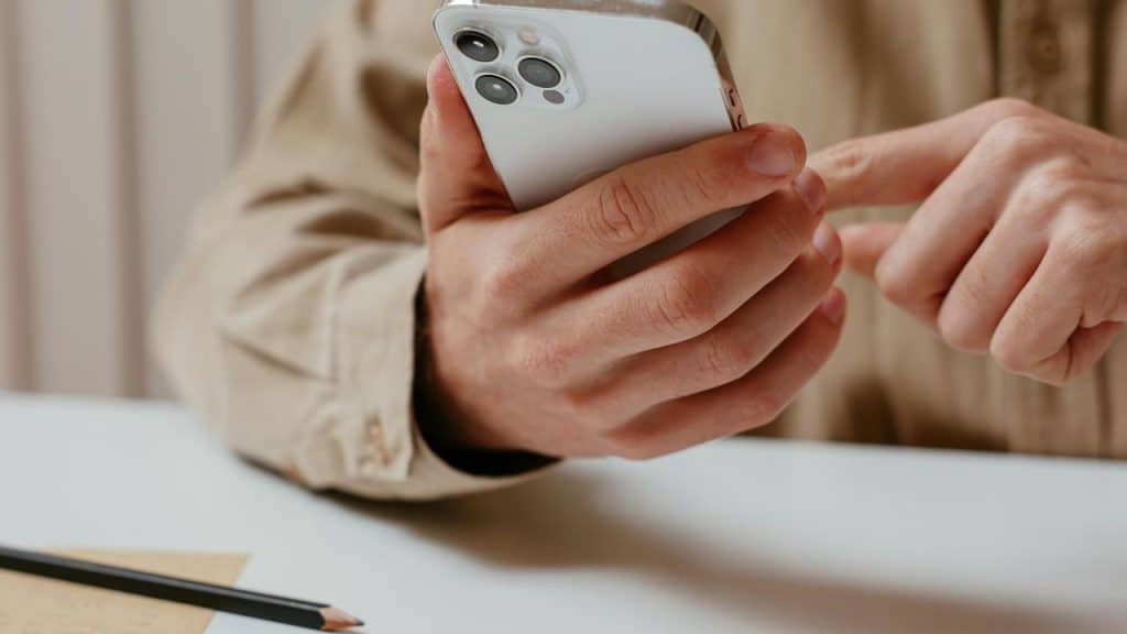 A person using a smartphone at a desk with a pencil and notes nearby.