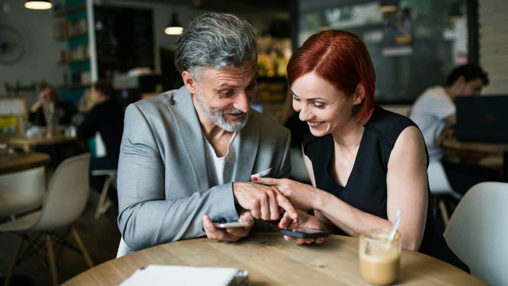 A mature couple smiling and looking at a smartphone together while sitting at a wooden table in a café.