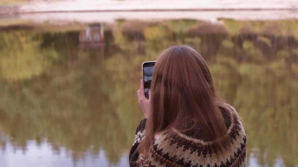 A woman standing by a lake taking a photo with her phone.