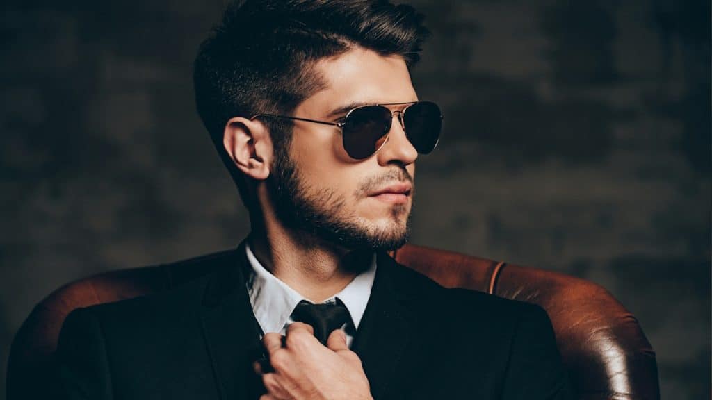 A stylish man in a black suit and aviator sunglasses sitting in a leather chair, looking away while adjusting his black tie.