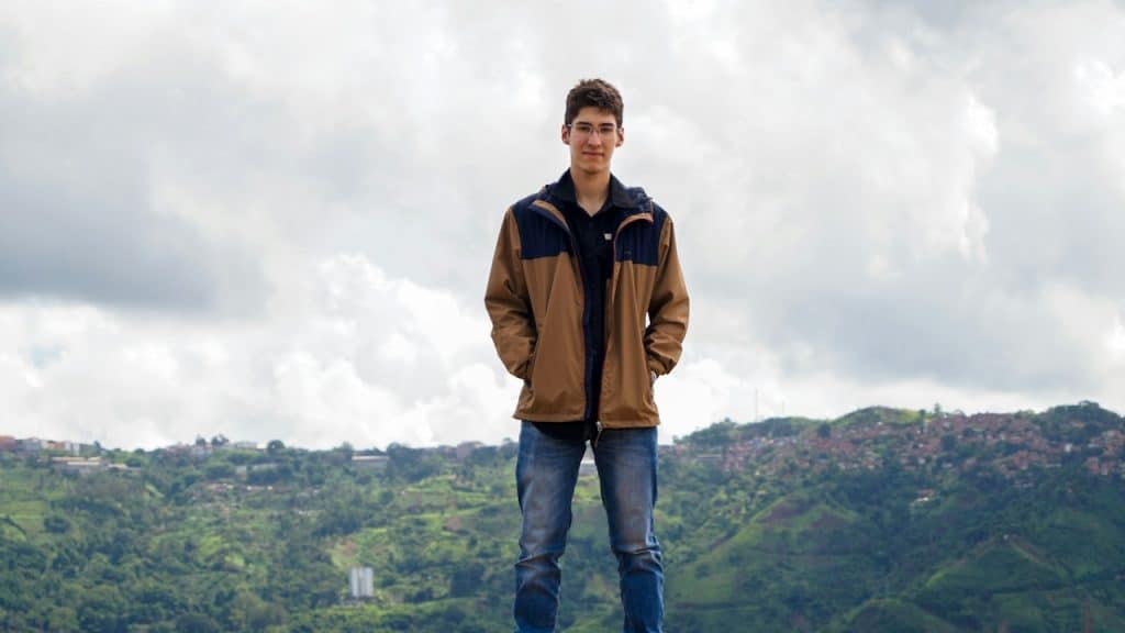 A young man in a brown jacket standing in a hilly area.