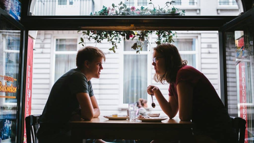 Silhouette of a young man and woman talking across a table inside a cafe, with the bright, blurred street view visible through the large window behind them.