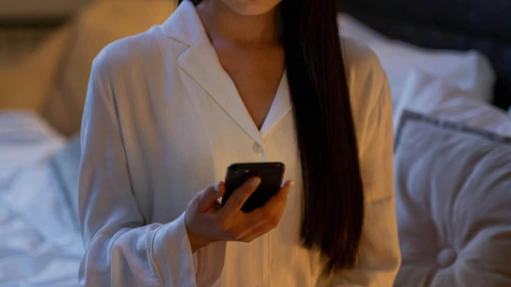 A person in pajamas holding a smartphone while standing by a bed.