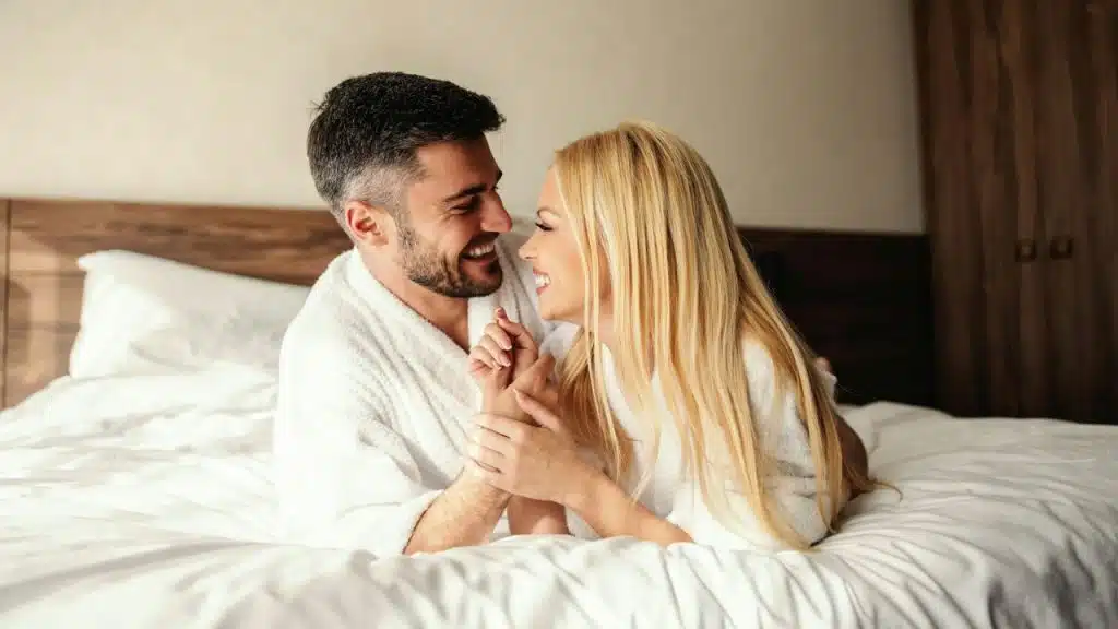 A smiling man and woman in white bathrobes are holding hands while lying in a bed.