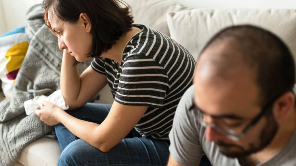 A distressed woman sits on a couch in a striped shirt holding a tissue, with a blurry man in the foreground.