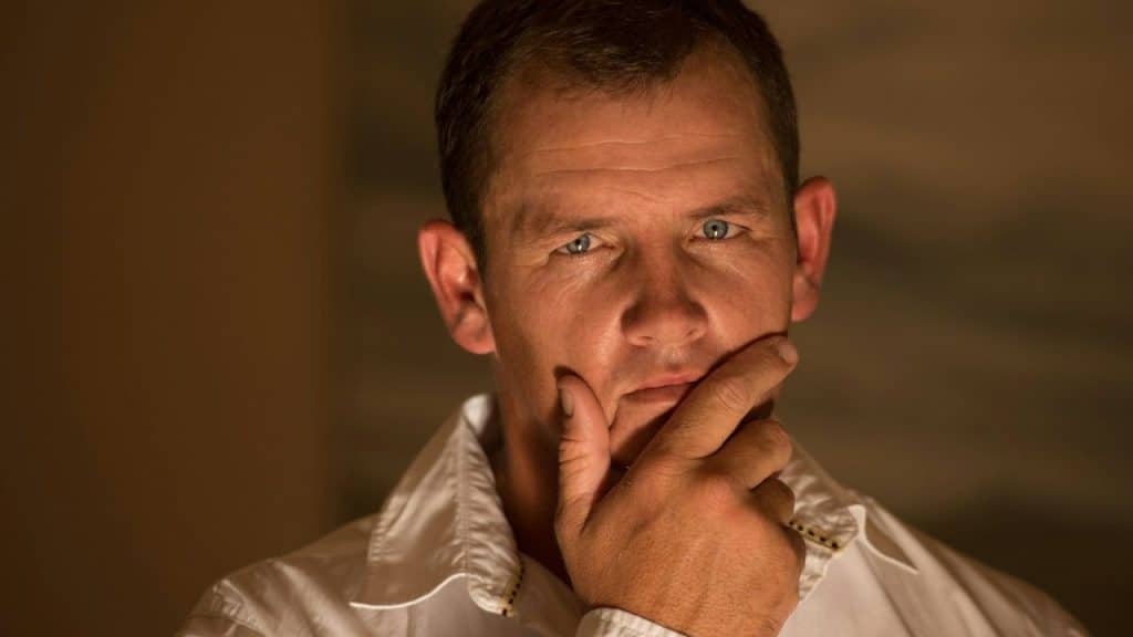 A middle-aged man is looking at the camera with his hand on his chin.