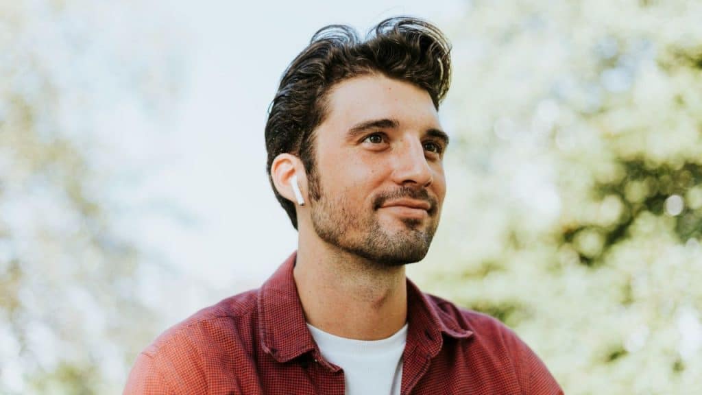 A smiling man wearing a red shirt and a white earbud is looking up outdoors.