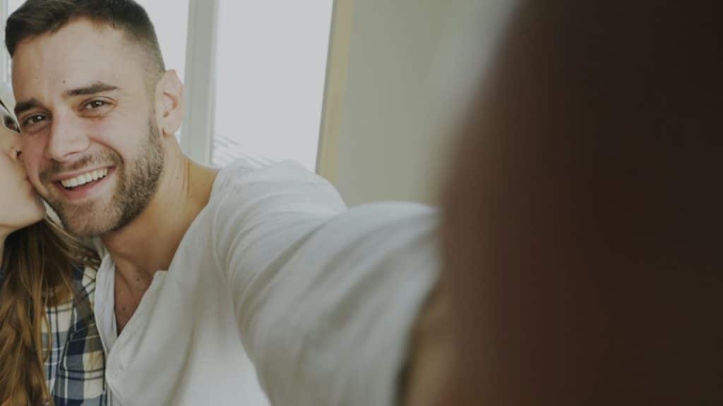 A smiling man takes a close-up selfie while a woman kisses his cheek.