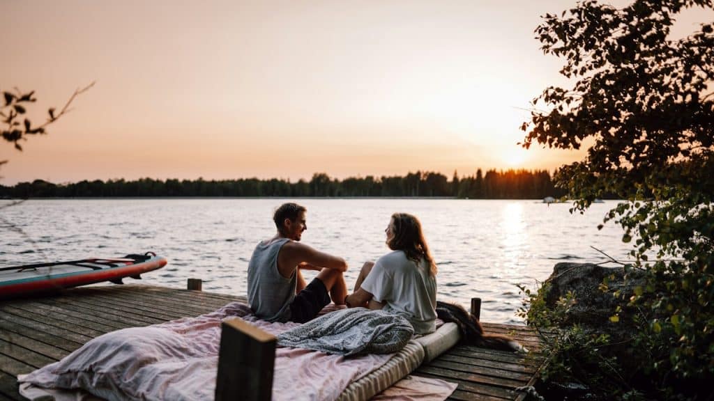 A couple sits on a mattress on a wooden dock, facing each other while watching the sunset over a lake.