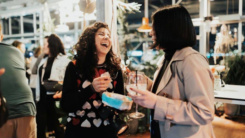 A woman laughing with a friend while holding drinks at a lively gathering.