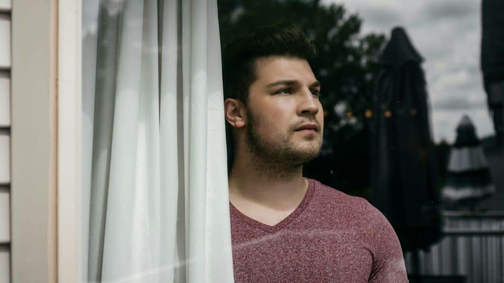 A man in a V-neck shirt looks out a window partially obscured by a curtain.