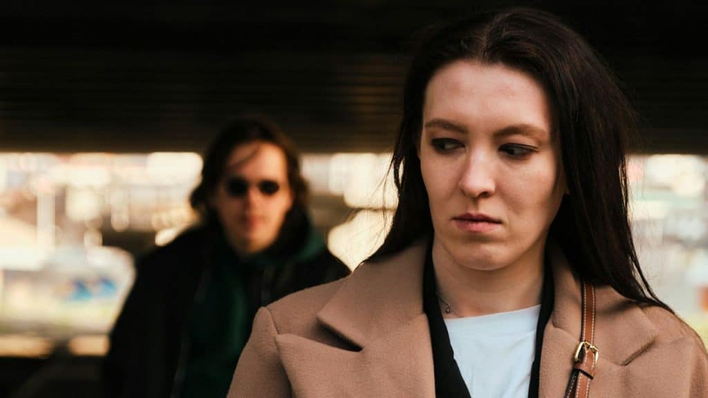 A somber woman in a beige coat looks down, with a blurred person in sunglasses behind her.
