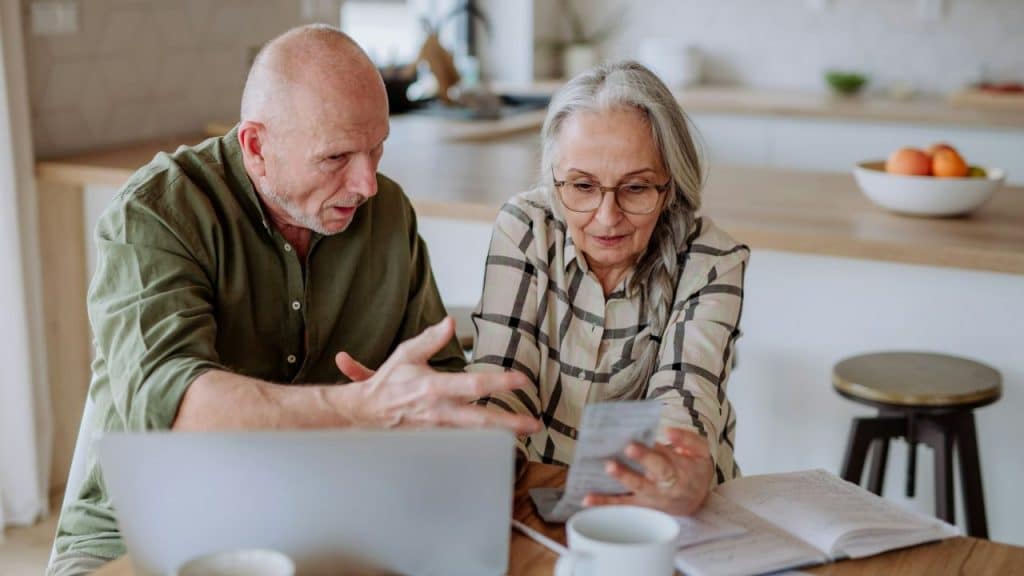 An older couple is sitting at a kitchen table, looking at papers and a laptop.