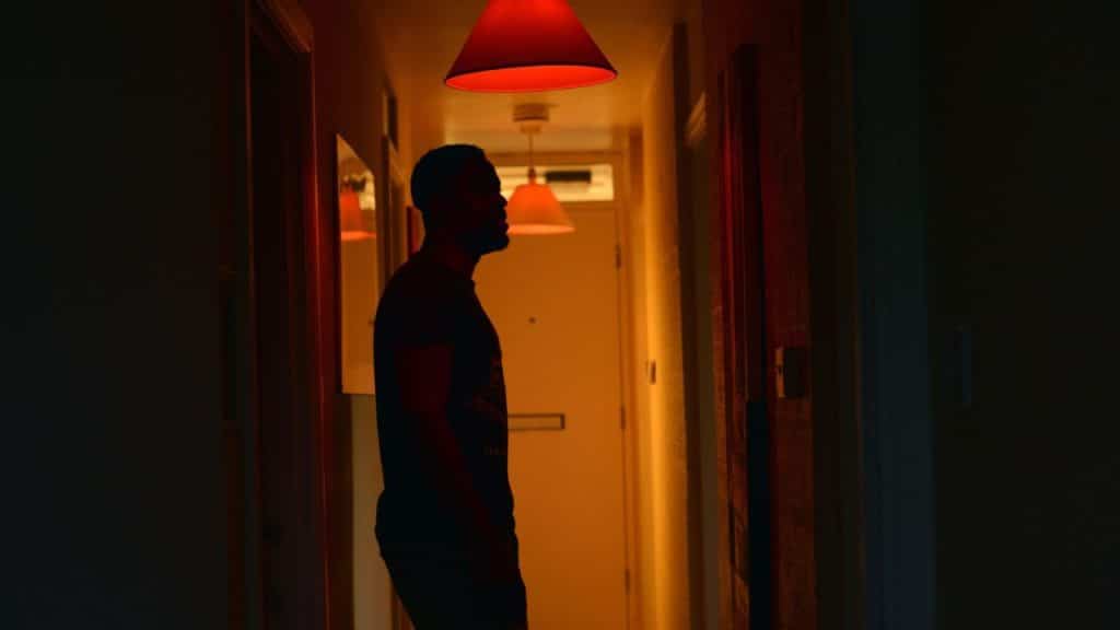 A silhouetted man stands in a dark hallway illuminated by a red overhead light.