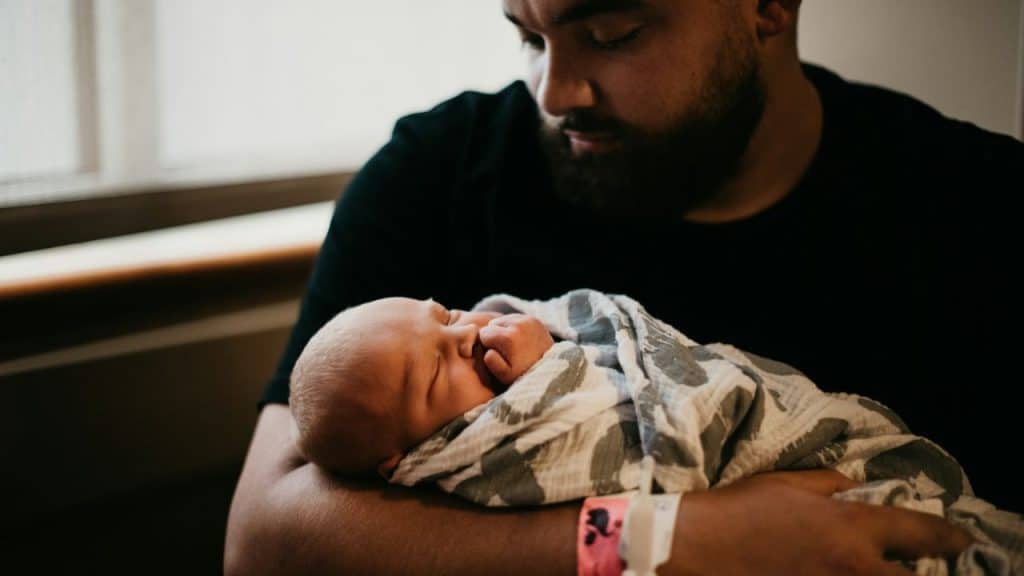 A bearded man in a black shirt holds a newborn baby wrapped in a patterned blanket.