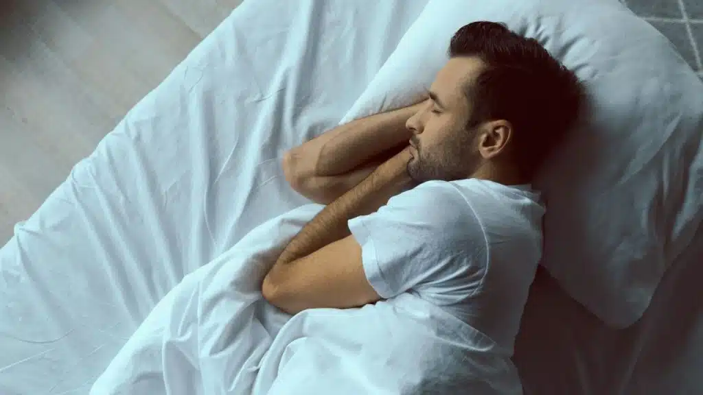 Man with a beard sleeping on his side in a bed with white bedding, arms tucked under his head.