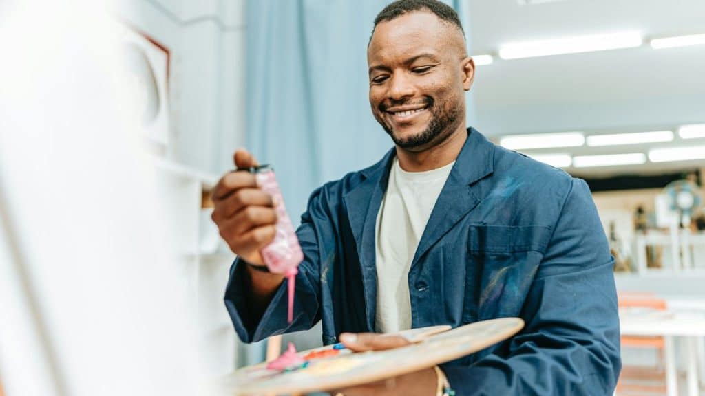 A smiling Black man is squeezing pink paint onto a palette in an art studio.