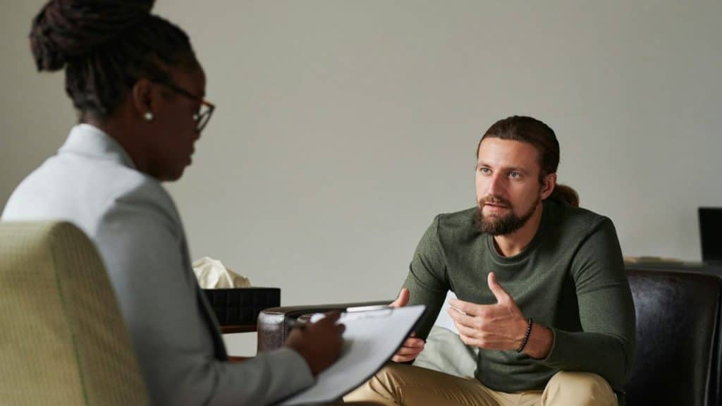 Man with a beard and long hair tied back, gesturing while speaking to a woman taking notes on a clipboard.