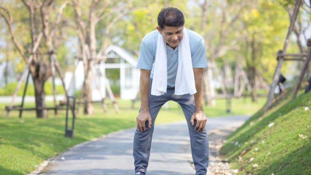 Man wearing a light blue shirt and a white towel around his neck, bent over and resting his hands on his knees in a park.