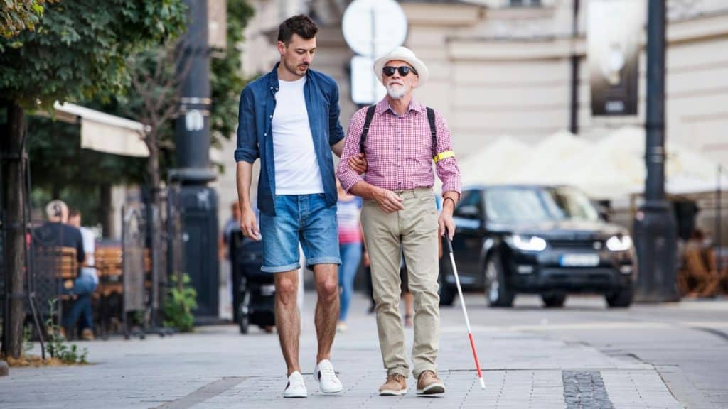 A young man is helping an older, visually impaired man walk on a city street.