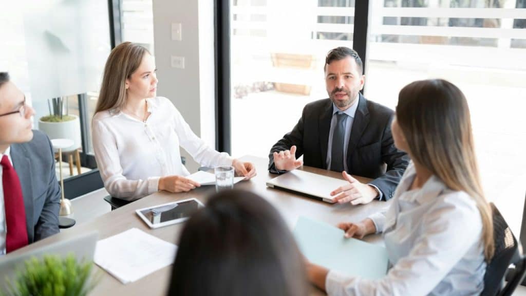 Four professionals meet at a conference table, with a man speaking and gesturing with his hands.