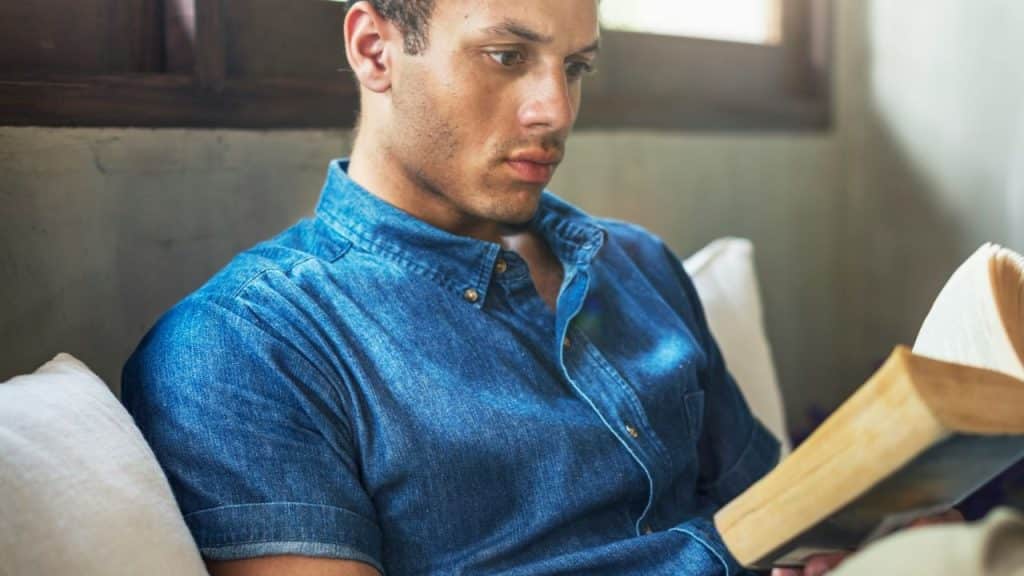 Man in a denim shirt sits indoors, intently reading a book with yellowish pages.