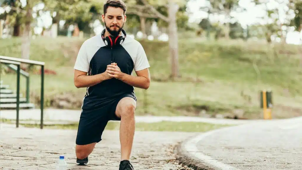 Man in athletic clothes and headphones performing a lunge exercise outdoors on a paved path.