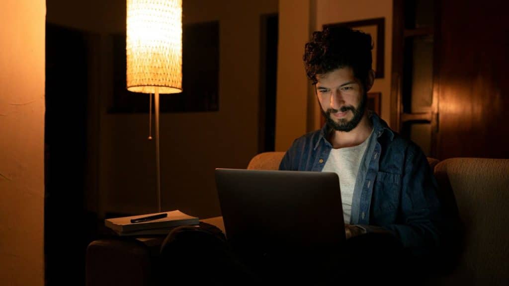 Man with a beard sits on a couch, focused intently on a laptop screen in the dark.