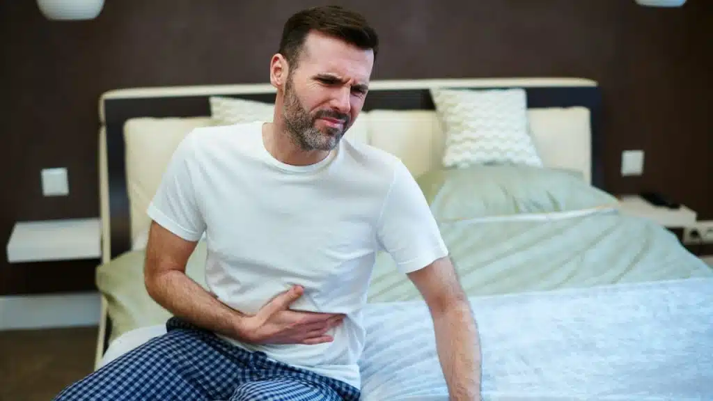 Man in a white t-shirt and plaid pajama bottoms sitting on a bed, clutching his side in pain.