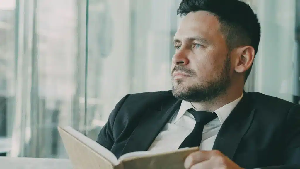 A serious man in a suit and tie sits with an open book, looking out a window.
