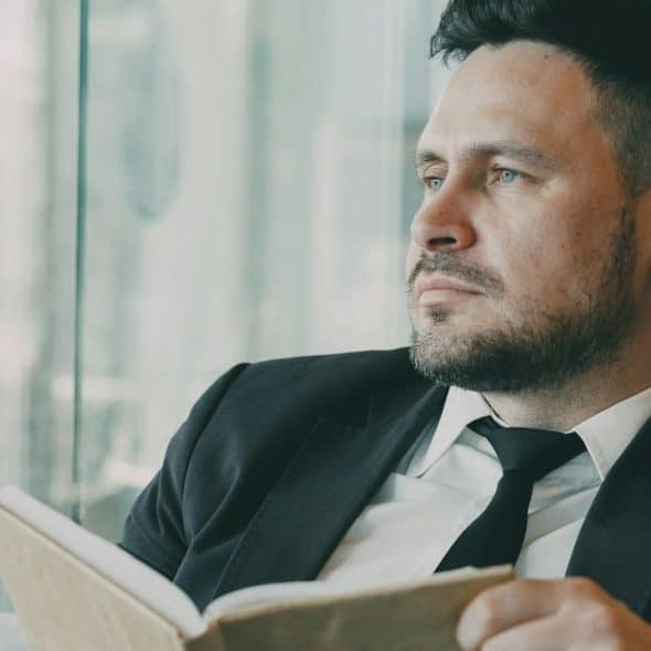 A serious man in a suit and tie sits with an open book, looking out a window.