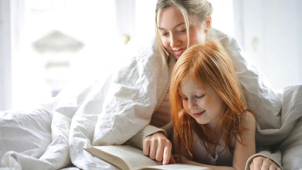 A mother reading a book with her daughter under a blanket.