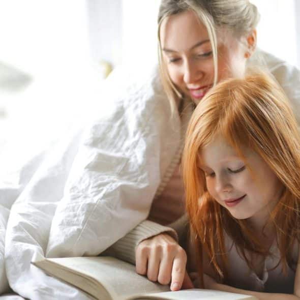 A mother reading a book with her daughter under a blanket.