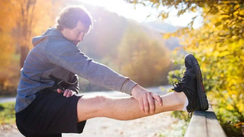 A man stretches his leg on a fence rail outdoors, with autumn trees in the background.