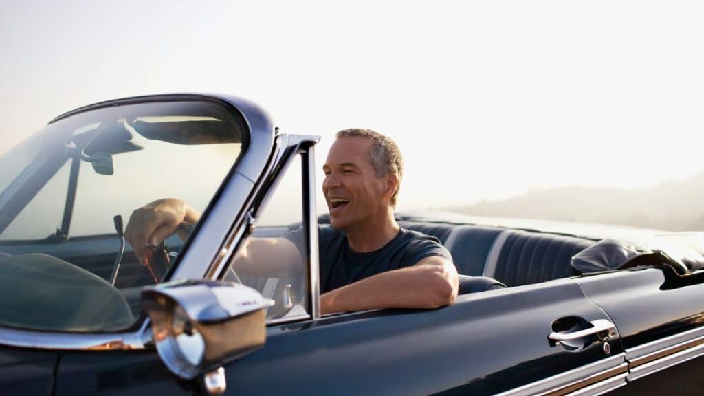 Smiling middle-aged man driving a dark-colored vintage convertible car with the top down.