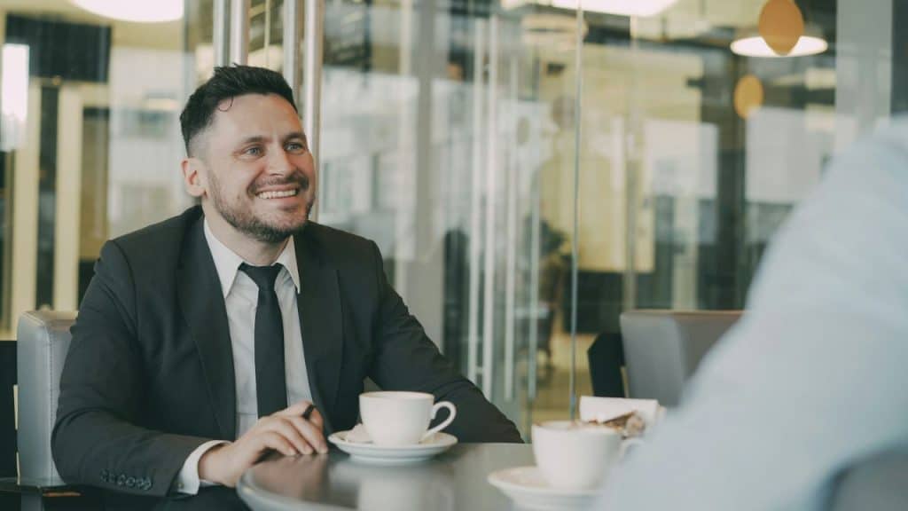 Man in a black suit and tie smiling during a conversation at a coffee table.