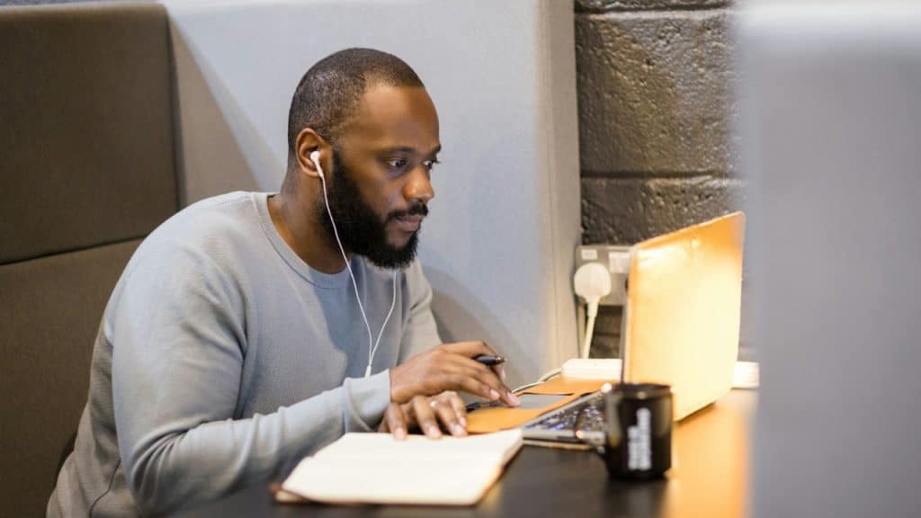 Bearded man with earbuds looking intently at a glowing laptop screen in a booth.