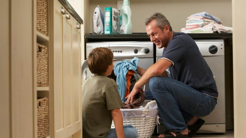 Man and boy crouching to put laundry into a washing machine in a laundry room.