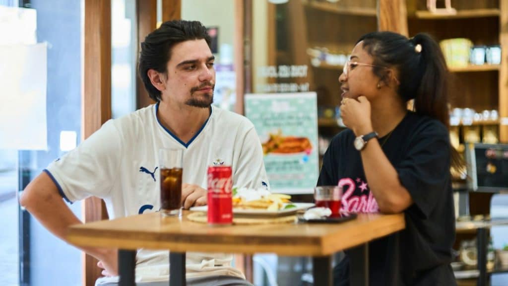 A young man and woman talking across a table with drinks and food in a cafe.