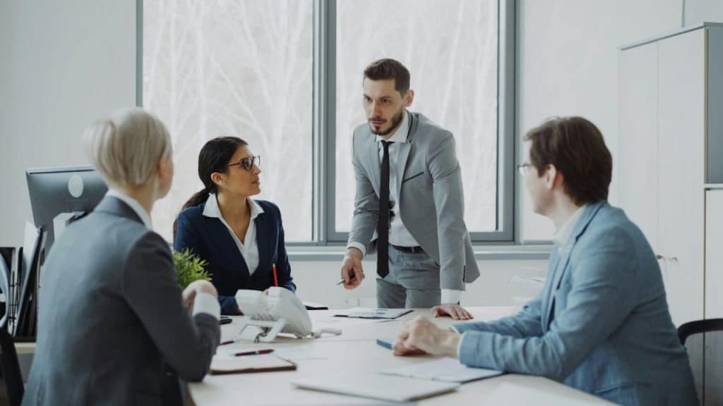 Man in a light gray suit standing at a conference table, addressing three people.