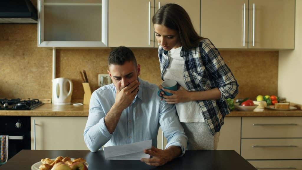 A concerned couple in a kitchen looking at a document, the man looking distressed.