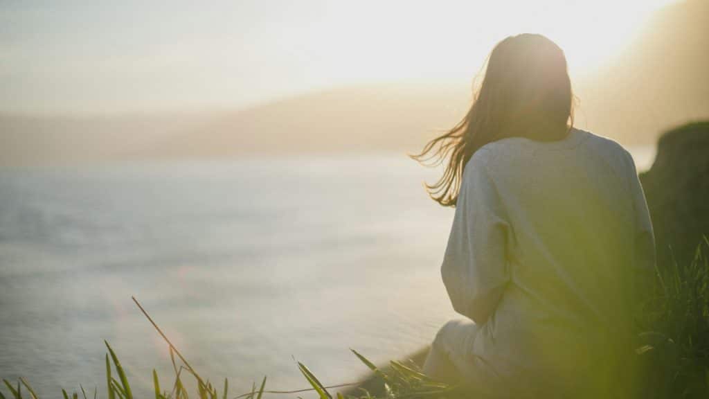 A woman sitting by the seaside with the sun shining in the background.