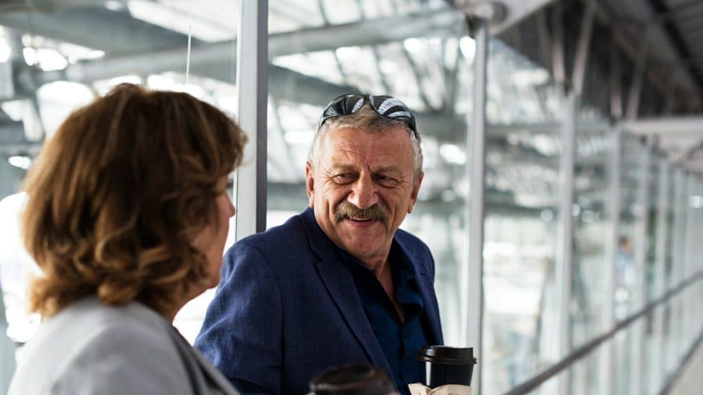An older couple engaging in a cheerful conversation indoors near a large glass railing, both holding takeout coffee cups.