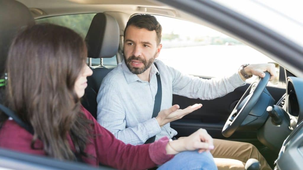 A man driving a car gestures while speaking to a woman sitting in the passenger seat.