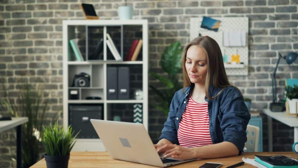 A woman with long brown hair sits at a desk, typing on a laptop in an office.